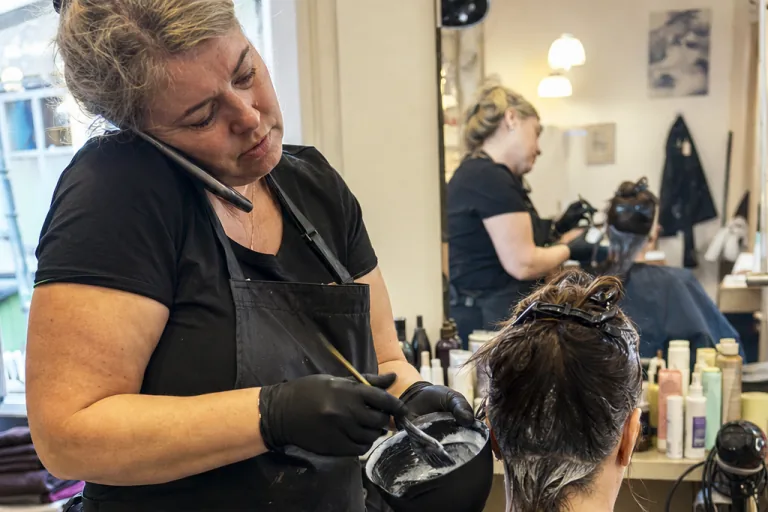 Friseurin klemmt das Telefon zwischen Ohr und Schulter, während sie mit Handschuhen, Farbschale und Pinsel an einer wartenden Kundin arbeitet.
